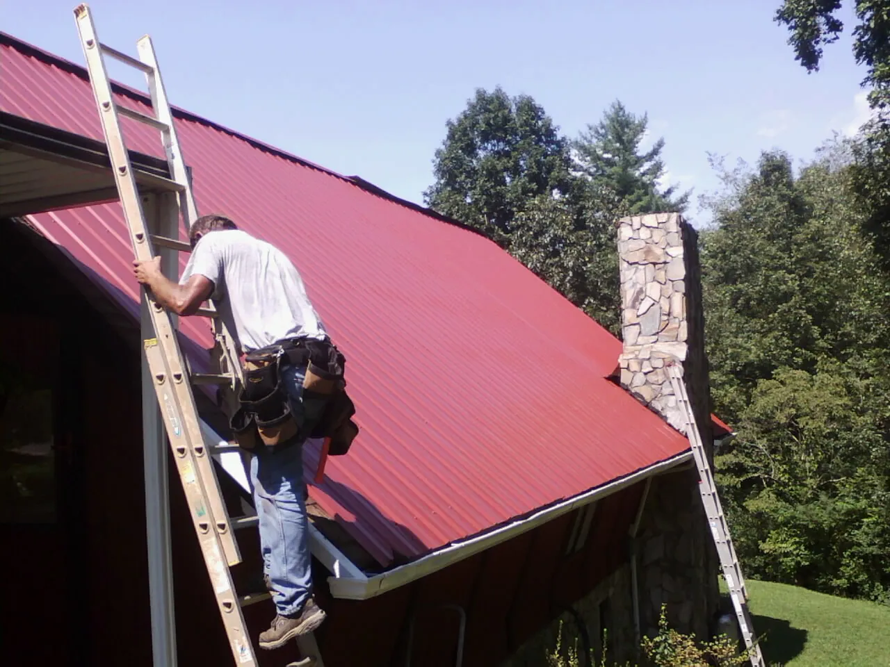 Roofer on a ladder beside a red metal roof and stone chimney — Soffit & Fascia Repair by Roofing Brothers in Falcon