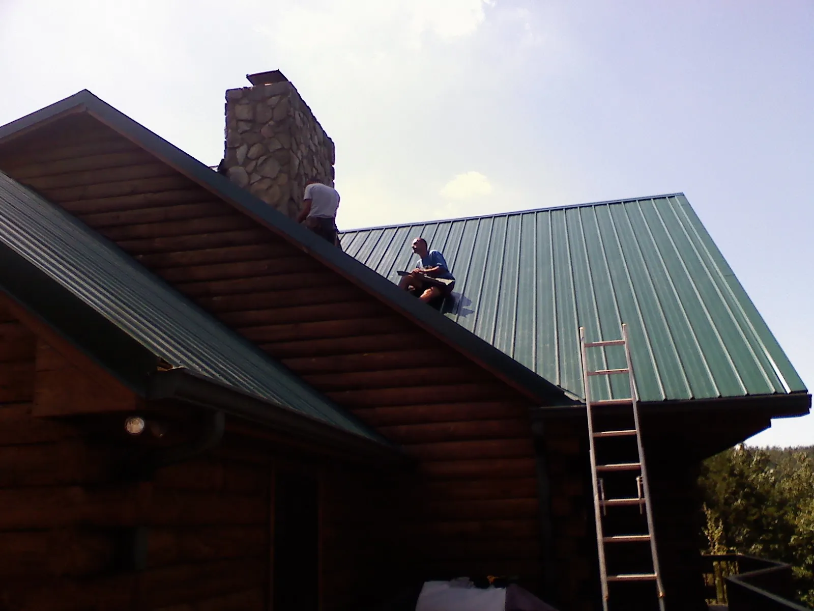 Roofers installing green metal panels beside a stone chimney on a log home in Falcon